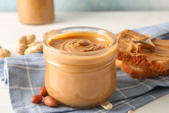 Glass jar with peanut butter, peanut, kitchen towel, spoon and peanut butter sandwich on wooden background, space for text and closeup