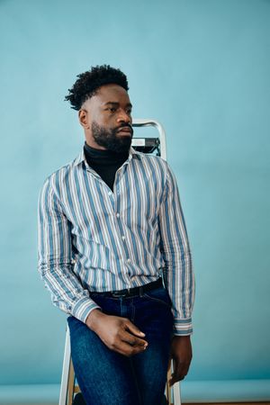 Somber Black male posing in blue studio in jeans and striped button up long sleeve
