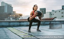 Female athlete doing squats holding a medicine ball standing on a rooftop 5lkxM0