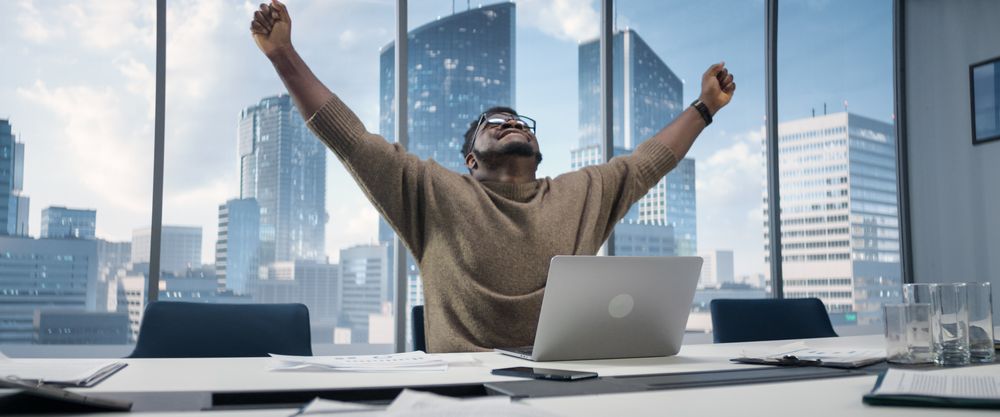 Black Businessman Sitting at His Desk in Skyscraper Office and Works on Laptop, Emotionally Celebrates Success in e-Business Deal.