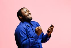 Happy excited successful modern man holding phone and raising arm up to celebrate achievement, isolated on pink background