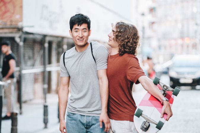 Cool multiracial male friends chatting while walking on street with longboard