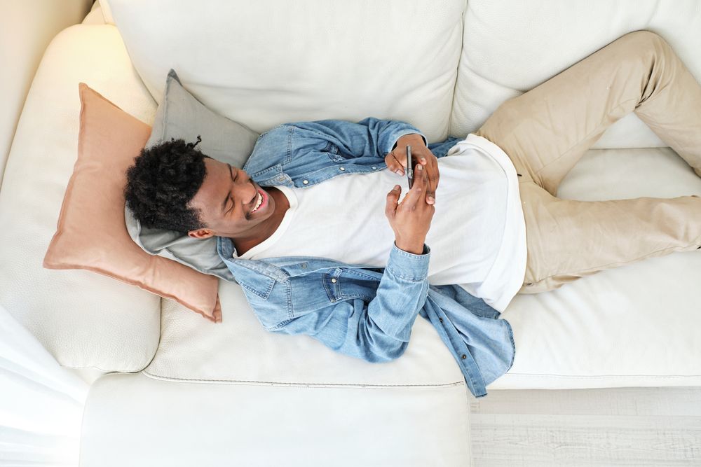 Happy Black man resting on sofa and using smartphone