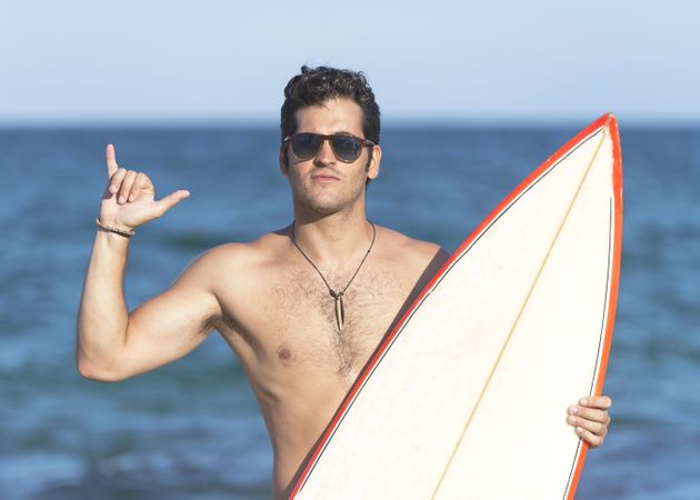 Male surfer standing with red outlined board near ocean making the “hang loose” sign