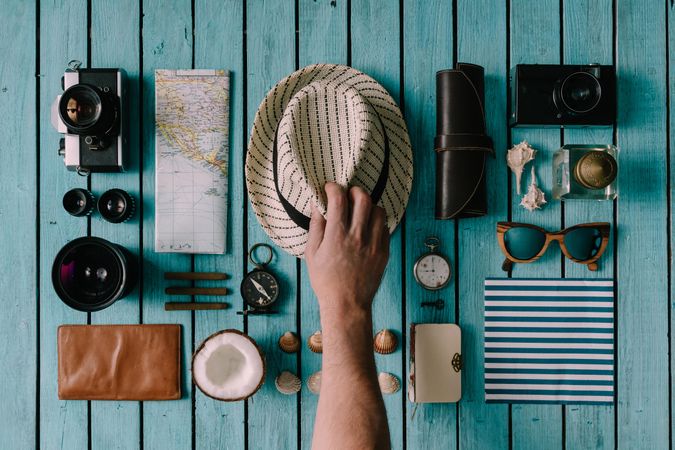 Man reaching for hat on neatly arranged travel items on blue background