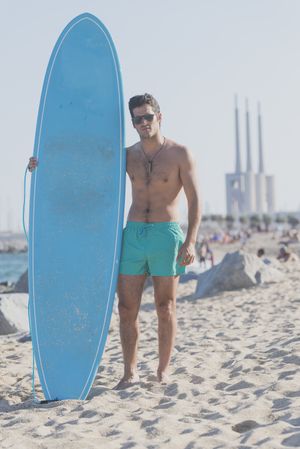 Full body of male surfer holding surfboard standing on the beach sand