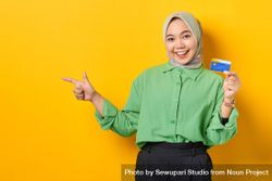 Smiling Muslim woman in headscarf and green blouse holding credit card and pointing aside 0VQ9rb