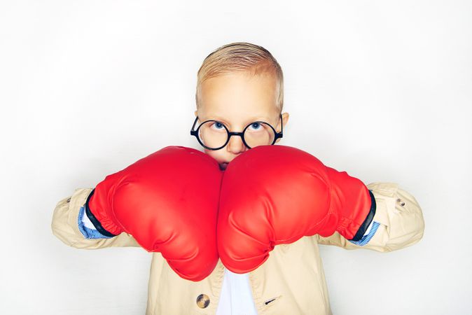 Serious blond boy wearing round glasses holding red boxing gloves together