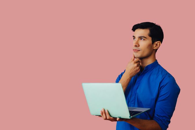 Contemplative Latino man holding laptop in pink room