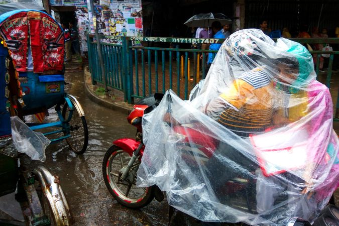 Man riding a motorbike with son covered with plastic sheet as rain shield in Dhaka, Bangladesh