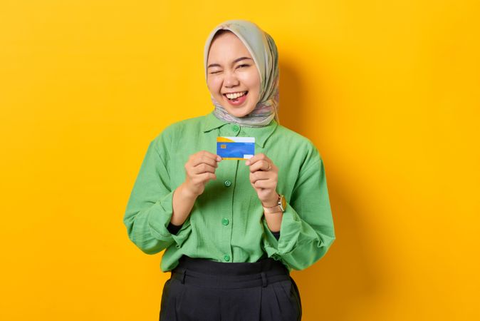 Muslim woman in headscarf and green blouse winking while holding credit card