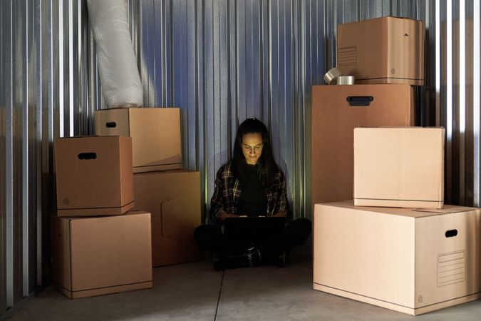 Young pretty woman inside a warehouse to store moving boxes organizing with her laptop