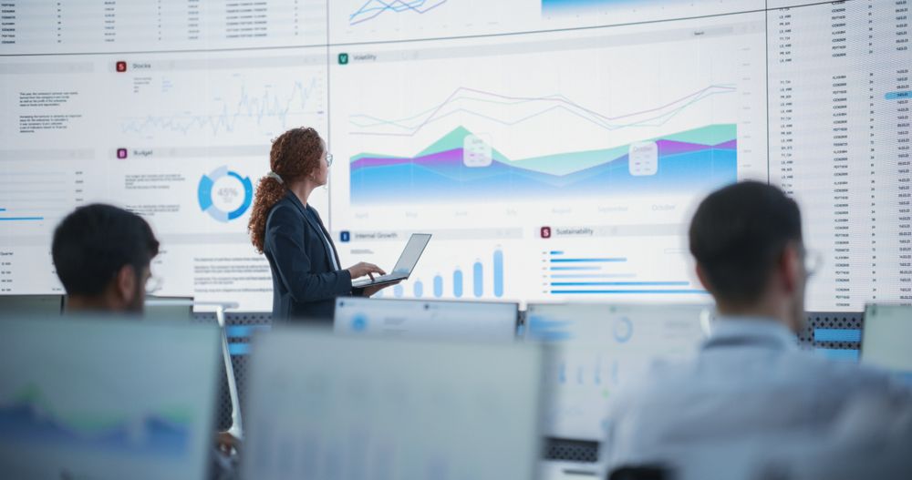 Female Data Scientist Using Laptop And Analyzing Information Graphs On Big Digital Screen In Monitoring Office.