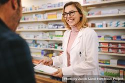 Smiling female pharmacist with prescription assisting a customer standing at the counter 0vqxBb