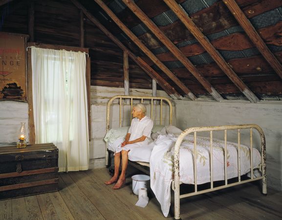 Kate Carter on her 90th birthday, sitting on bed for shoot in log cabins, North Carolin