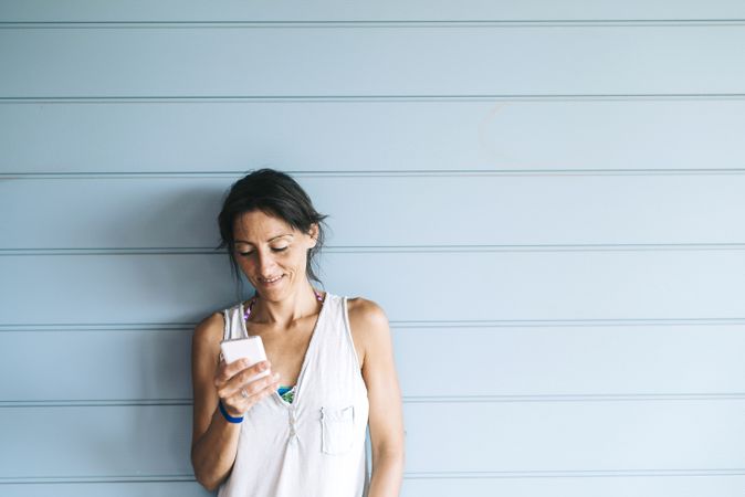 Calm woman in summer dress checking phone while leaning on wood wall