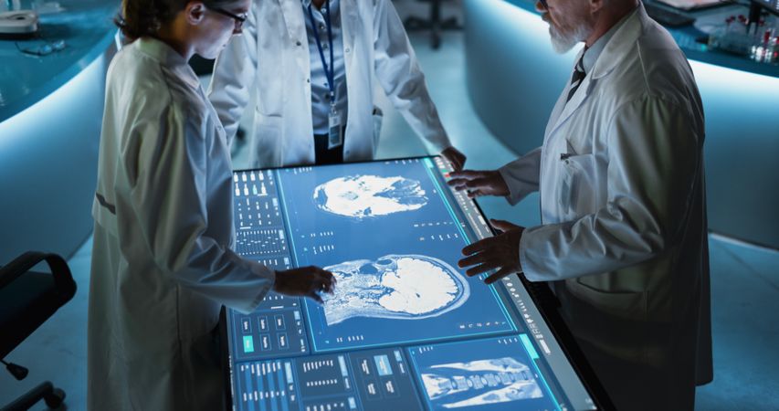 Modern Hospital Medical Research Center: Diverse Colleagues Gathered Around Interactive Touch Screen Table With CT Scans Of Brain On Display.