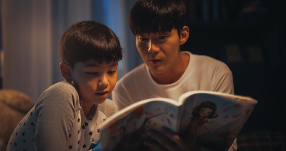 Korean Father Reading a Fairytale to His Lovely Cute Son in Bed Before Going to Sleep.