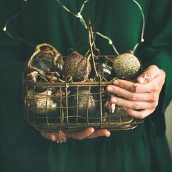 Woman in green dress holding wire basket of gold holiday decorations, square crop