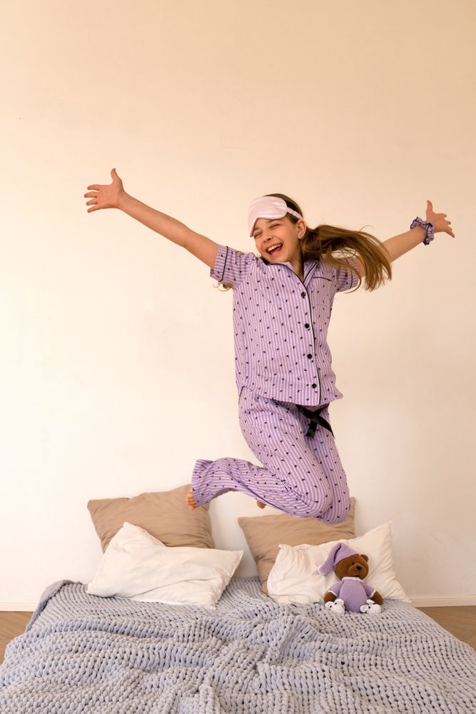 Joyful girl jumping on bed in purple pajamas.