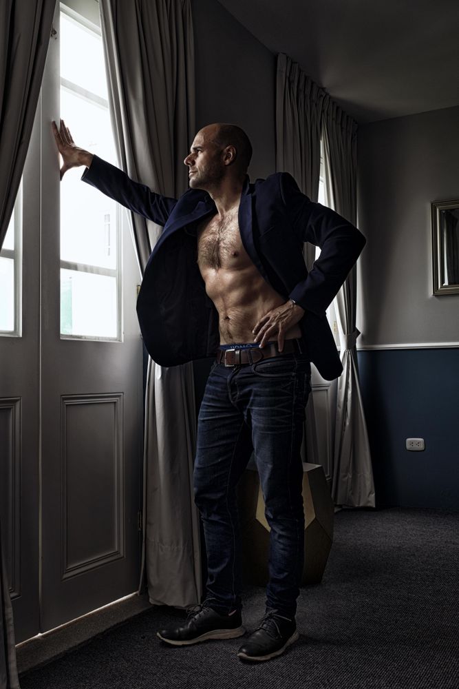 Bare chested man in dark blazer standing beside window