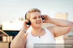 Blonde woman in exercise gear holding headphones to her ears outside bxmeM0