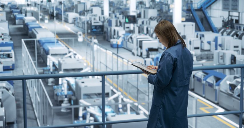 Thoughtful Asian Engineer Monitoring and Analyzing Conditions at a Pristine Modern Electronics Factory with Automated Robot Hands Working with the Help of Artificial Intelligence Software