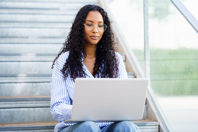 A woman is working on her laptop while comfortably sitting on outdoor steps in sunlight