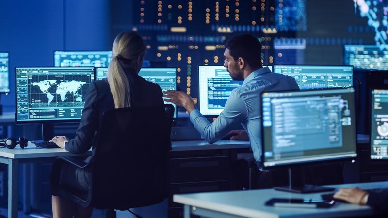 Two Professionals Collaborate in a High-tech Control Room, Analyzing Cybersecurity Data on Multiple Screens. the Environment Promotes Strategic Planning and Innovative Solutions.
