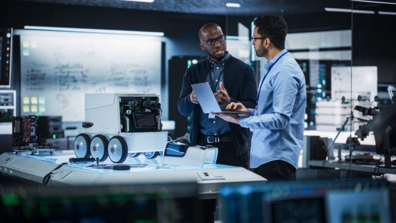 Portrait of Two Diverse Male Engineers Developing a Remotely Controlled Robot in an Industrial Research Facility. Specialists Using Laptop Computer, Working on Machine Improvement, Brainstorming