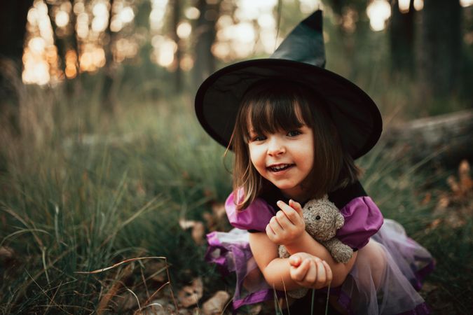 Girl in witch costume crouched down in the forest smiling with her teddy bear