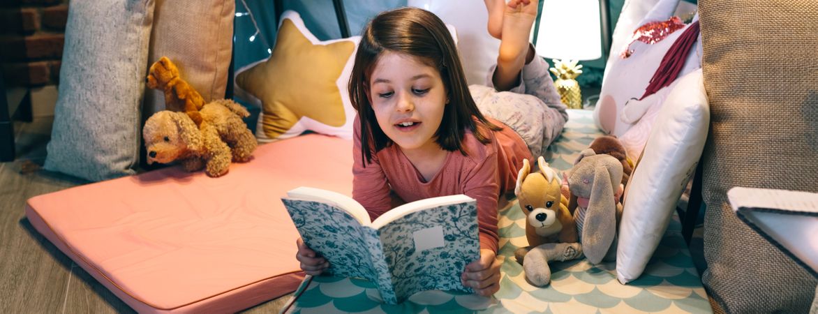 Banner of happy little girl reading and relaxing lying over sleeping mat in a cozy tent at home