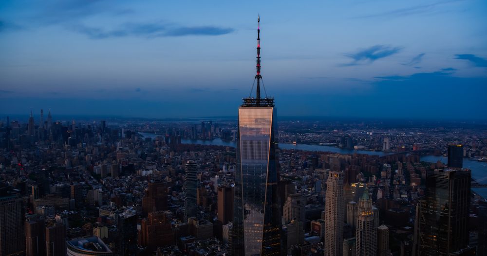 Late Evening Aerial Shot of a Camera Locked on Top of the One World Trade Center Skyscraper with Antenna. Helicopter Flying Around the Glass Building with a View on All Five New York City Boroughs