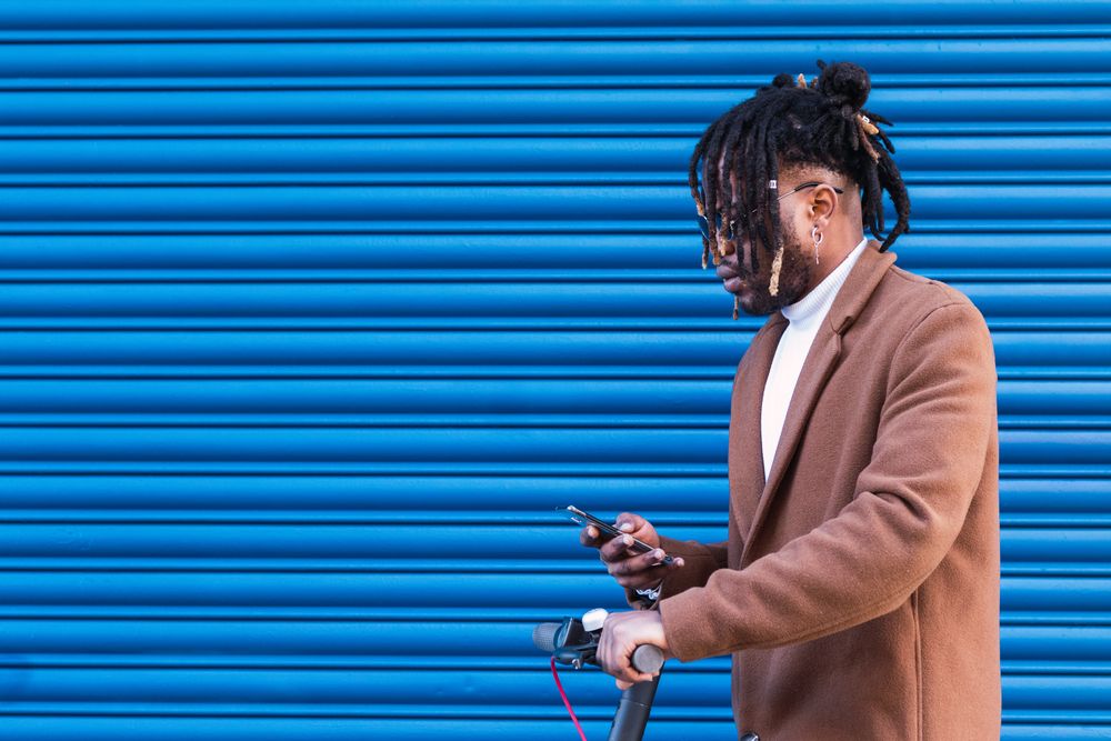 Stylish Black man with electric scooter using smartphone
