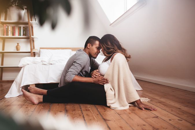Romantic young couple sitting on floor of bedroom holding mother’s belly