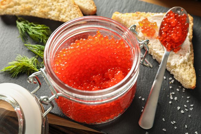 Composition with caviar in glass jar, bread, spoon and cutting board, closeup
