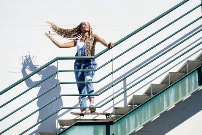 Joyful female in denim overalls standing on skateboard on stairs playing with her hair