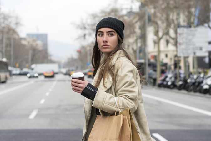Portrait of female in hat crossing the street, with takeaway coffee and shopping bag in winter
