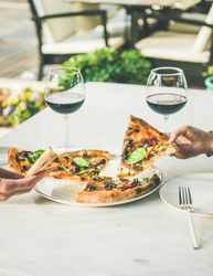Two people taking opposite slices of freshly made pizza with red wine