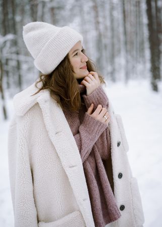 Woman in light knit cap and coat standing on snow covered ground in woods