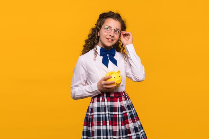 Girl adjusting glasses while holding yellow piggy bank.