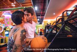Couple kissing each other standing in a gaming parlour holding basketballs 4Oo1R4