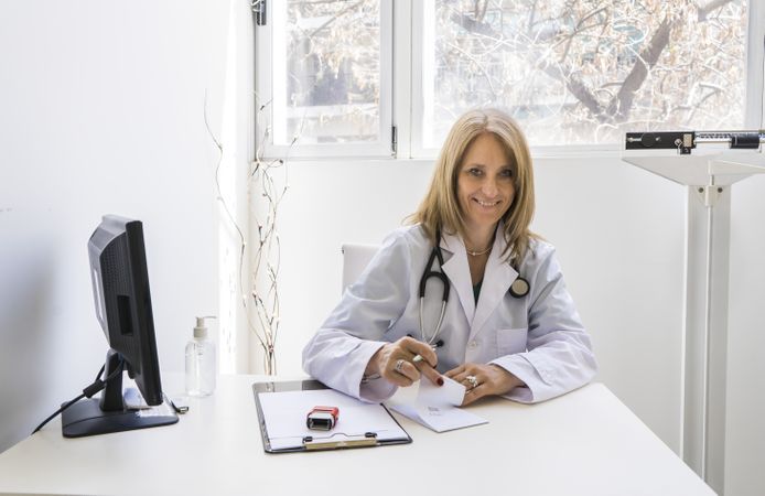 female doctor is writing a prescription at her medical office while looking at camera