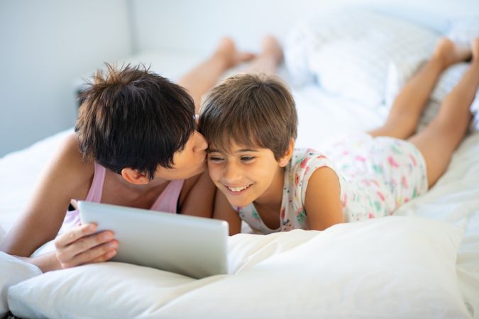 Woman kissing daughter’s forehead in bed as she watched tablet