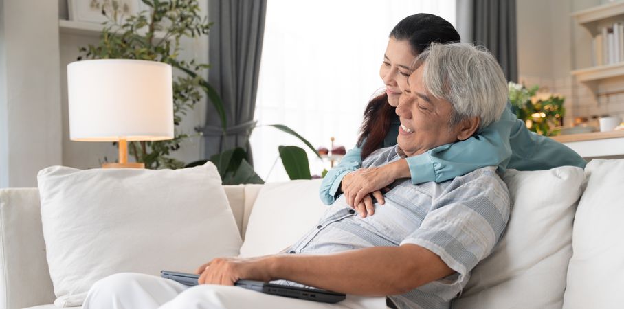 Asian mature man sitting on couch working on laptop while his wife embraces
