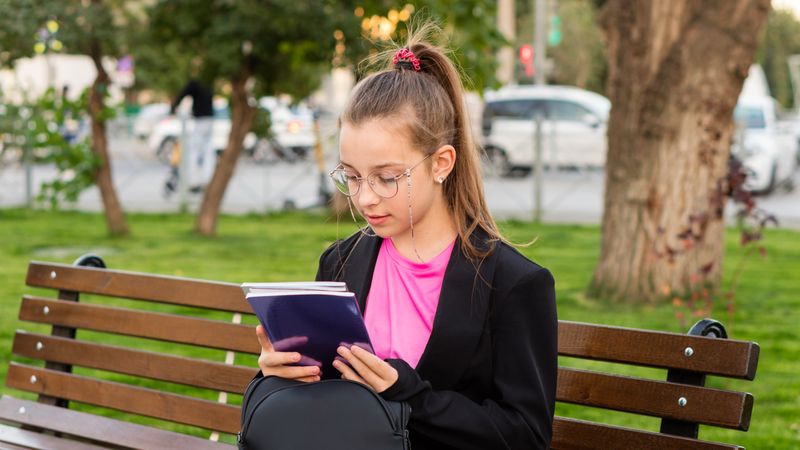 Girl reading book while sitting on park bench.