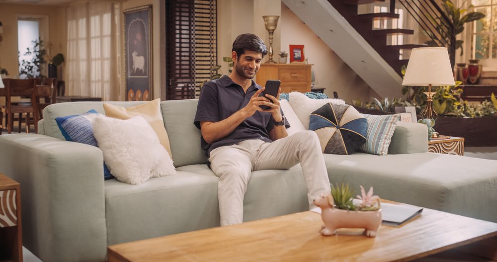 Cheerful Young Hindu Man Comfortably Using His Smartphone At Home, Handsome, Smiling and Positive