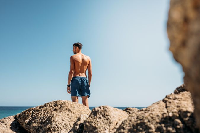 Rear view of a shirtless man standing on top of rocks near the sea