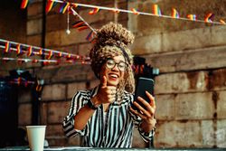 Smiling Black woman in stripe shirt giving thumbs up to phone in cafe