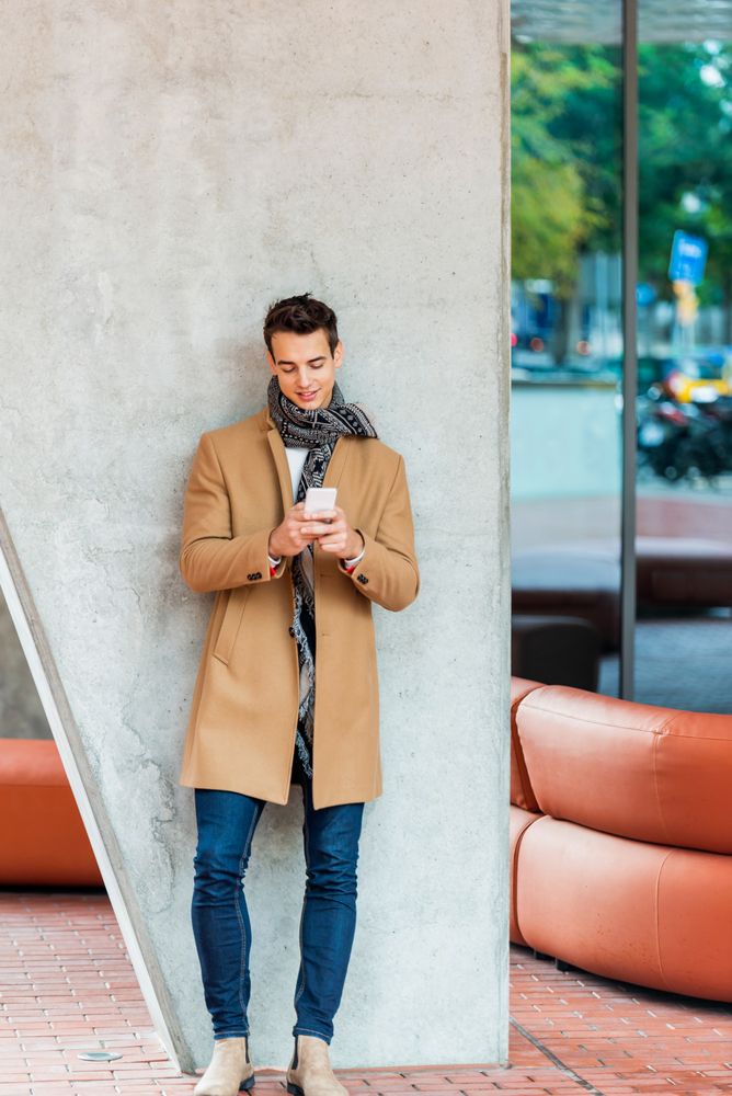 Coy man leaning on wall outside texting on phone and wearing a camel coat 
